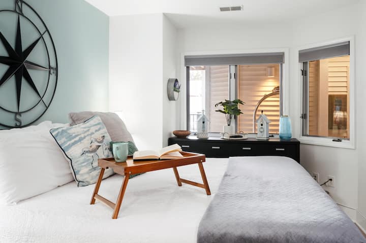 Relax in this bright, modern bedroom featuring a cozy breakfast tray, soft coastal accents, and plenty of natural light for a perfect start to your day. ☕