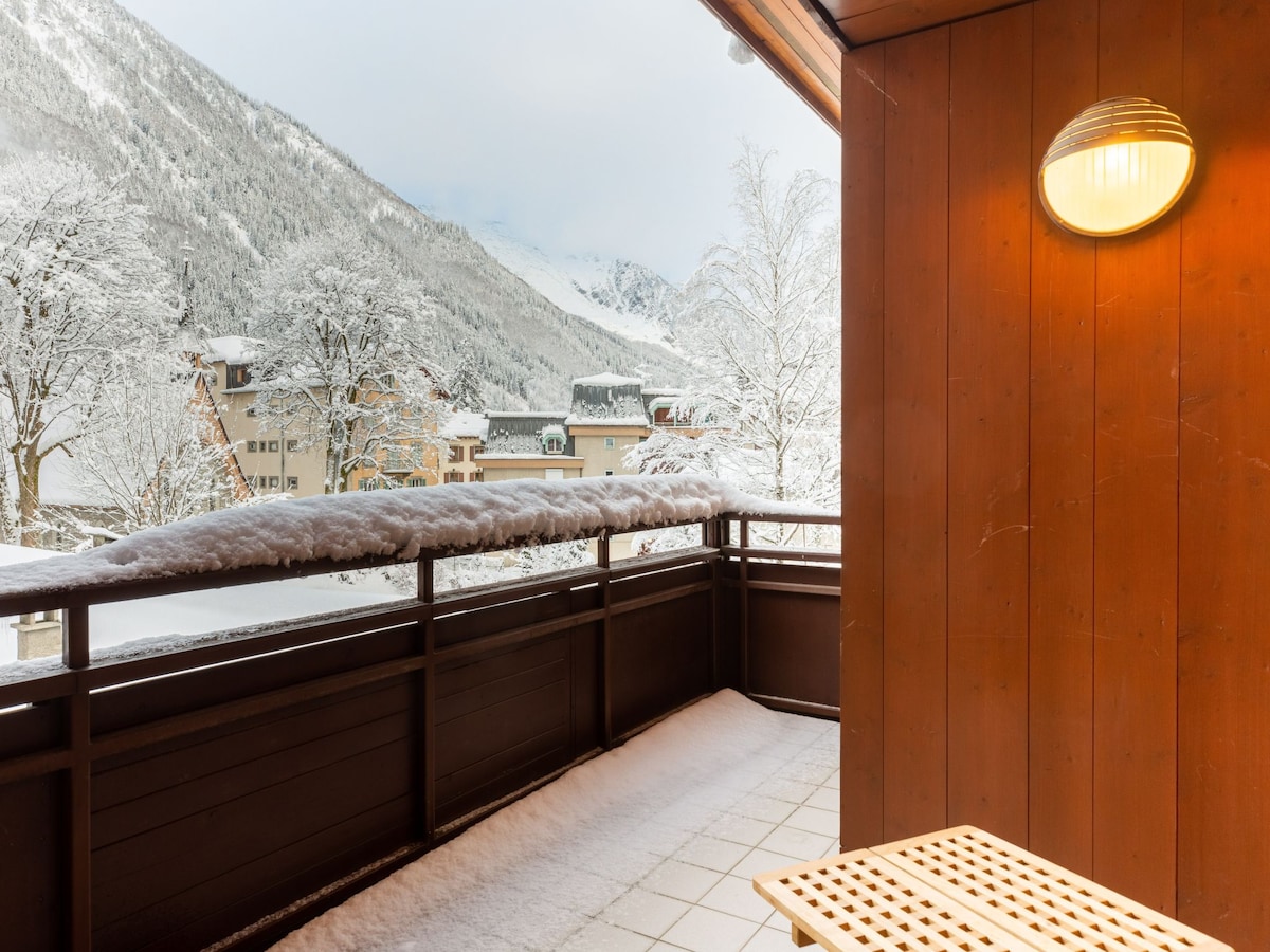 A balcony is shown, featuring a wooden bench positioned beside a railing. Snow blankets the floor and surrounding landscape, with a panoramic view of the snow-covered mountains in the distance and the buildings below.