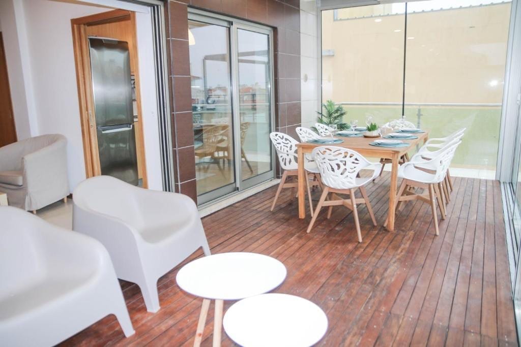An outdoor dining area features a large wooden table surrounded by eight white chairs, ideal for gatherings. Two small white side tables are positioned on a wooden deck. Large glass doors provide access to the interior space, with views of the surroundings.