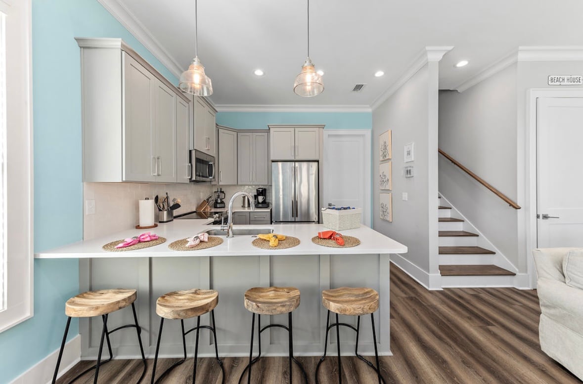 A modern kitchen area features a large island with white countertops and four wooden stools arranged around it. Stylish pendant lights hang above, illuminating the space. Natural light enters through nearby windows, while stainless steel appliances are visible in the background, complemented by light cabinetry and soft blue walls.