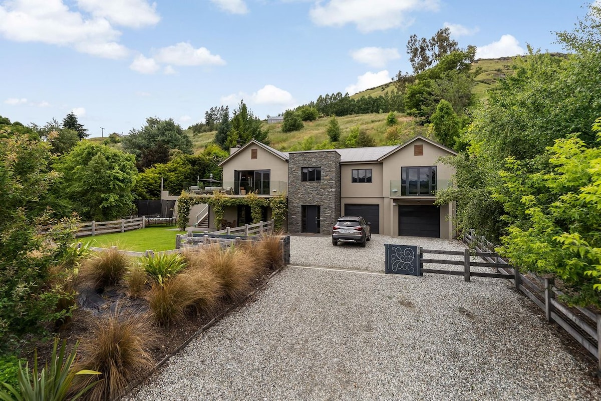 A lodge-inspired home is set amidst lush greenery, with a gravel driveway leading to the front. The two-story structure features a stone facade, spacious garage, and well-maintained lawn. Surrounding trees enhance the natural setting, offering a sense of privacy.