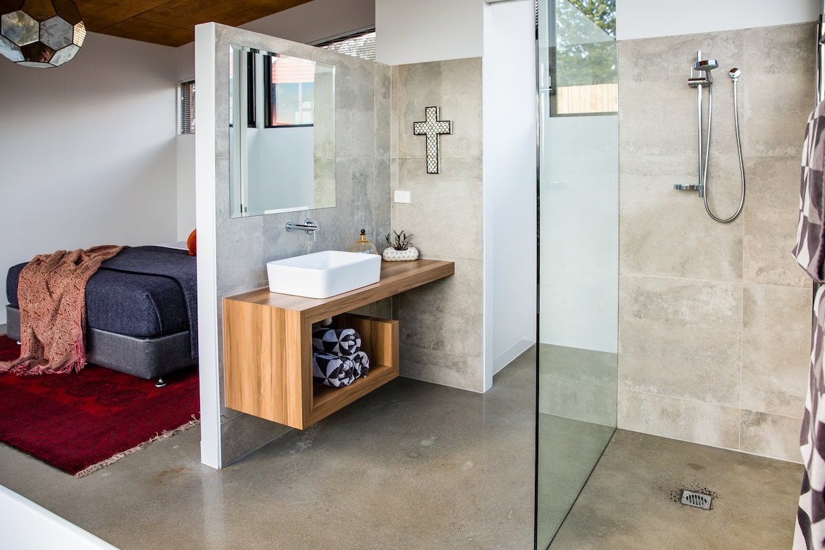 An airy bathroom is designed with a modern shower enclosure and sleek wooden vanity. A spacious mirror above the sink reflects the room's neutral tones. A cozy bedroom area is visible in the background, featuring a bed with a decorative throw and a soft rug.