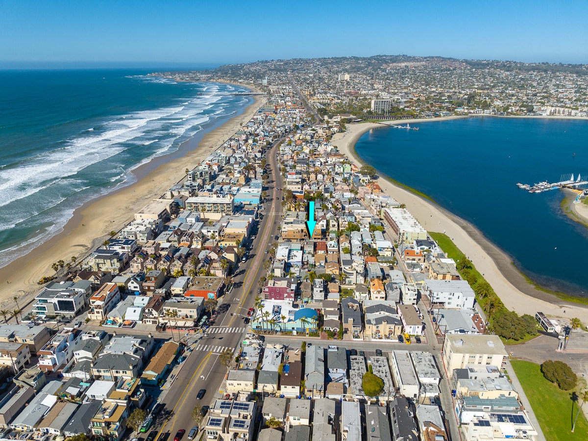 An aerial view captures a coastal neighborhood, showcasing a blend of residential buildings and proximity to sandy beaches and a bay. Waves can be seen breaking along the shoreline, while the vibrant layout includes streets, houses, and green areas, illustrating the inviting atmosphere of the area.