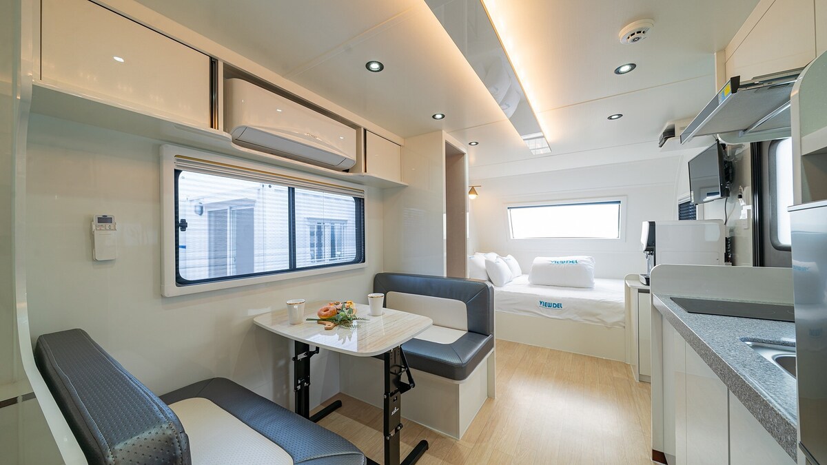 A clean and modern space features a combined living and sleeping area. A spacious bed is positioned against the wall, while a dining table with seating for four is adjacent, illuminated by natural light from the large window.