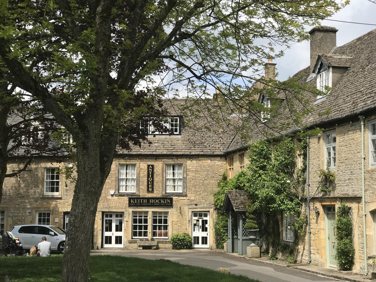 A charming exterior view of a historic building features local Cotswold stone architecture. The entryway is framed by a blooming tree and manicured lawn, while the inviting façades showcase traditional windows and a sign reading 'KEITH HOCKIN.' Natural light enhances the warm tones of the stonework.