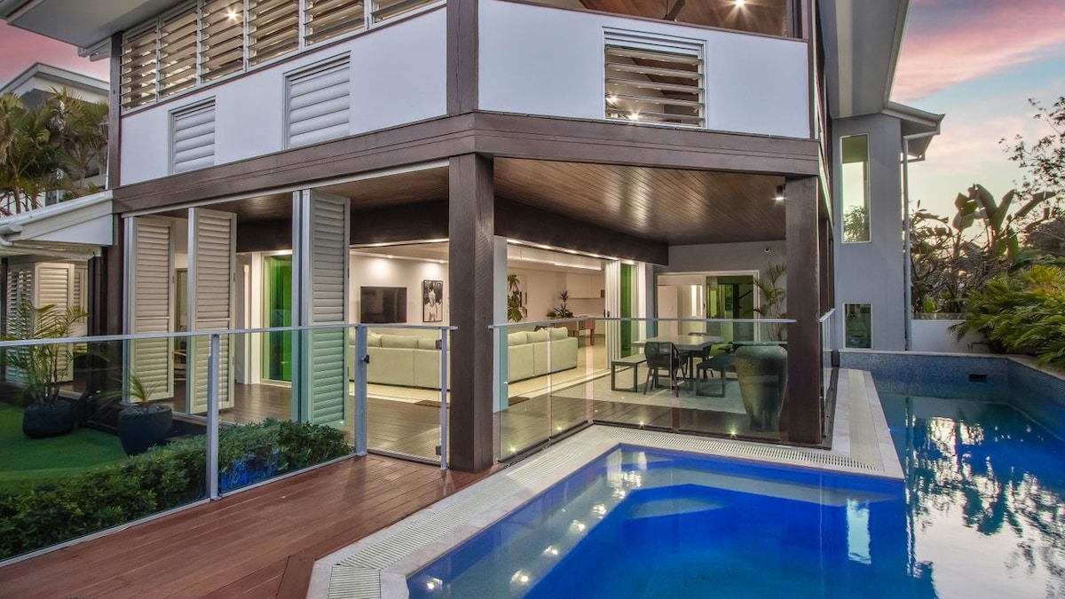 The image displays a modern two-story home featuring expansive glass doors that open to a private pool area. The solar-heated pool is surrounded by lush greenery. The indoor dining and living spaces are visible through the open design, promoting seamless indoor-outdoor living.