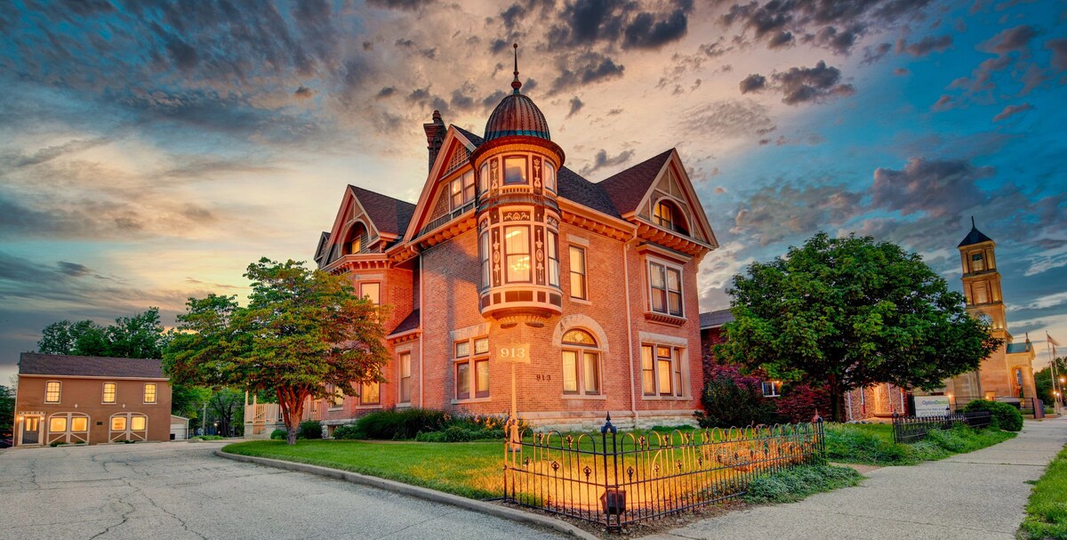 The exterior of a historic Queen Anne-style Victorian house is illuminated by warm evening light. The structure features intricate architectural details, including a prominent turret and decorative trim. Lush landscaping surrounds the home, with a wrought iron fence outlining the property.