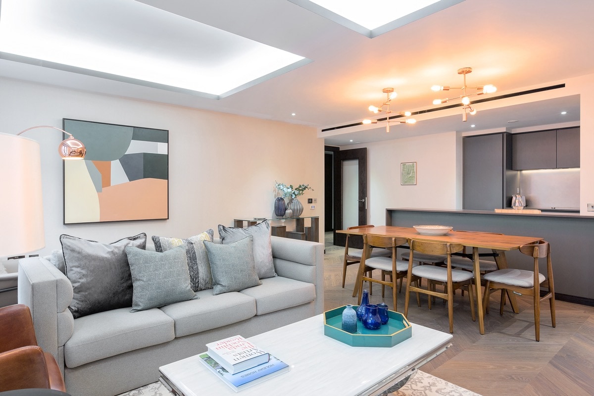 A modern living area is furnished with a light gray sofa adorned with plush cushions. A sleek coffee table sits in front, with books and decorative items displayed. A dining table with matching chairs is visible in the background, complemented by warm lighting and a stylishly designed kitchen area.