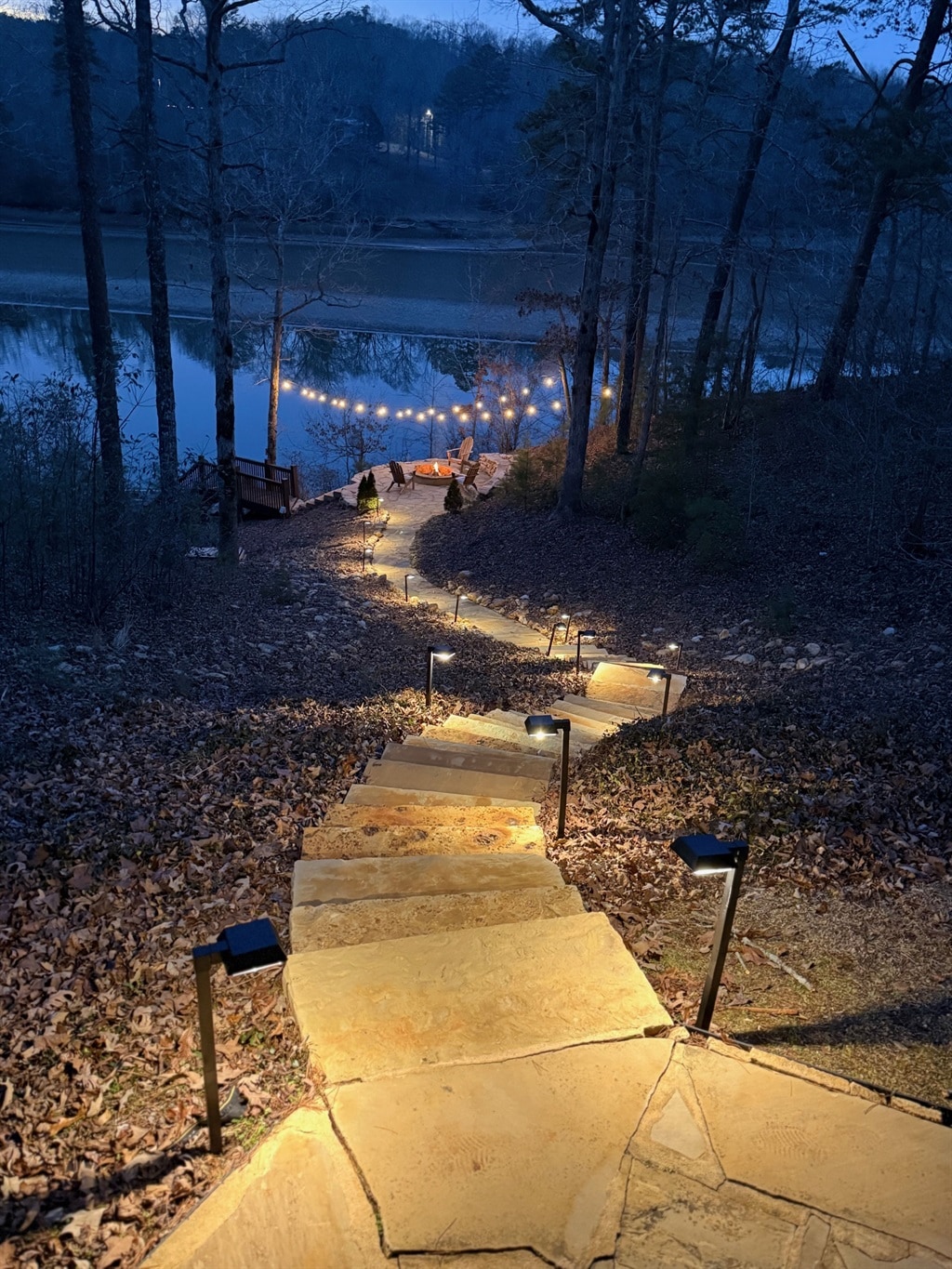 A stone pathway is lined with soft lighting, guiding downwards through a wooded area towards a picturesque lake. The tranquil water reflects the evening ambiance, while a cozy seating area with string lights is visible at the water's edge, inviting relaxation.
