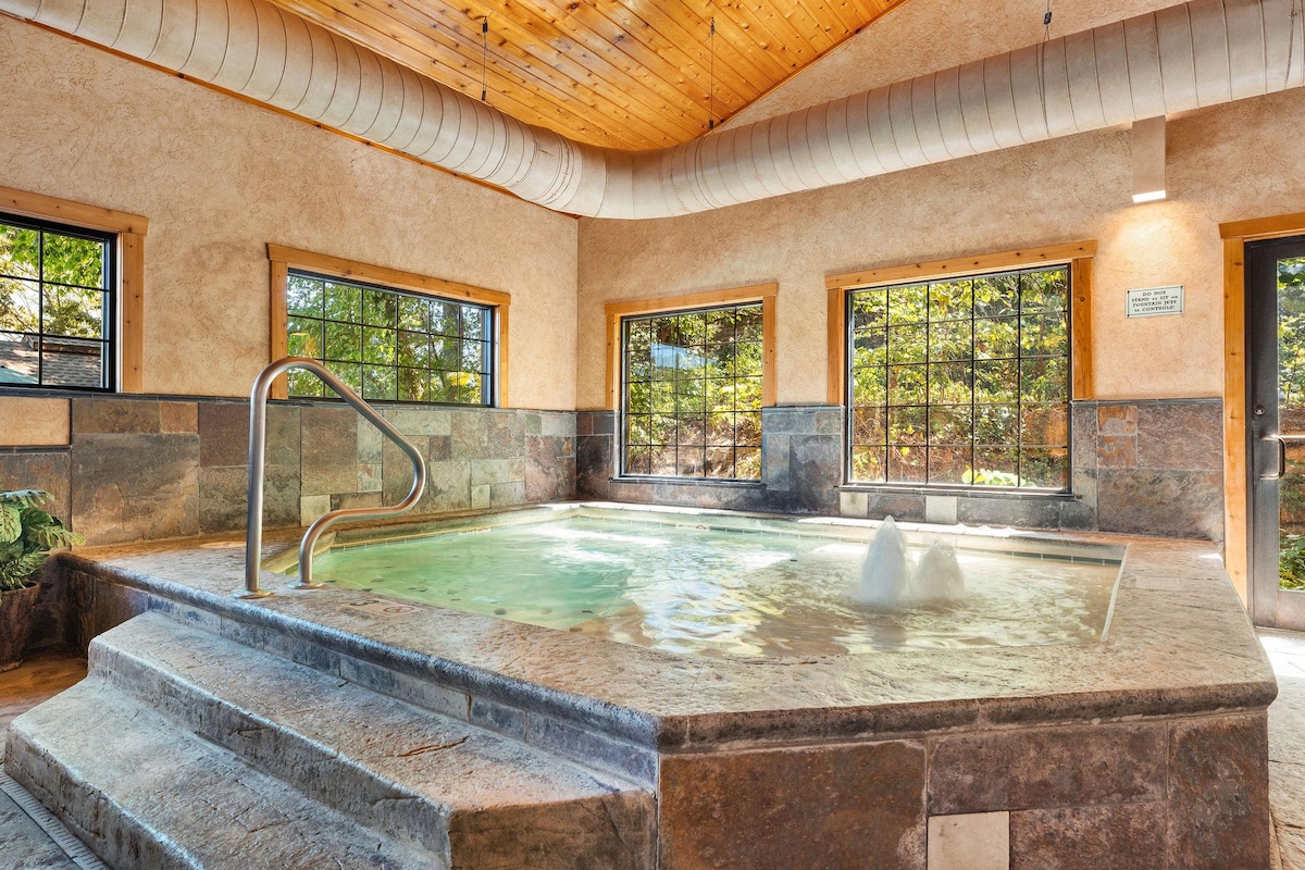 An inviting Jacuzzi is situated in a room with high ceilings and large windows showcasing natural light. The surrounding stone walls enhance the serene ambiance, and a set of steps leads into the warm water, providing easy access.
