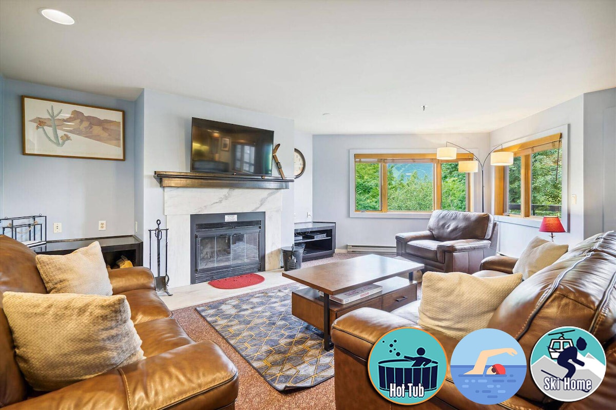 The spacious living room features a stone fireplace with a flat-screen TV mounted above it. Large windows allow natural light to fill the space, highlighting the comfortable brown leather sofas arranged around a wooden coffee table.