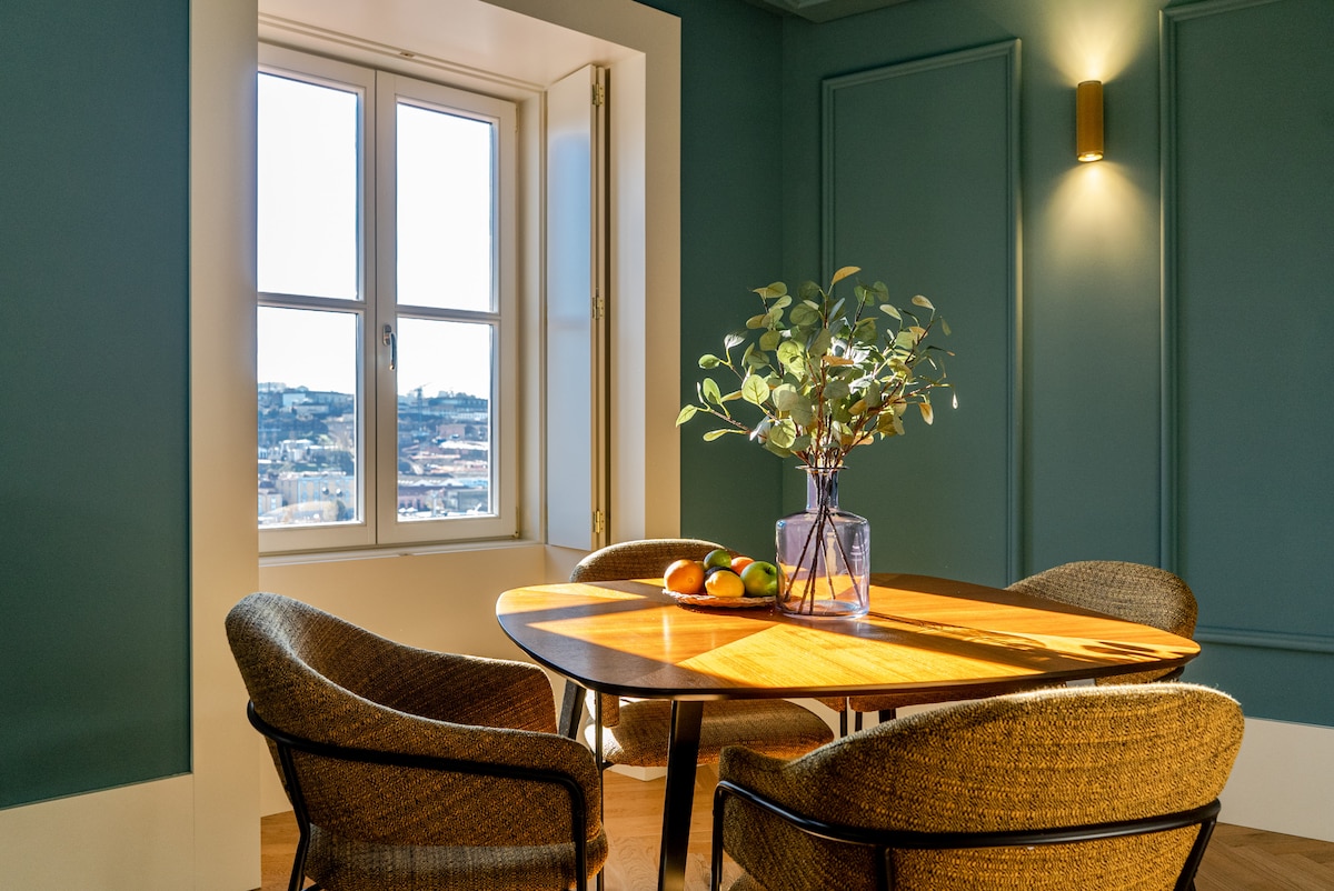 A dining area features a round wooden table surrounded by four woven chairs. A floral arrangement is placed in a clear vase at the center of the table. Natural light streams through a large window, illuminating the space and revealing a distant city view.