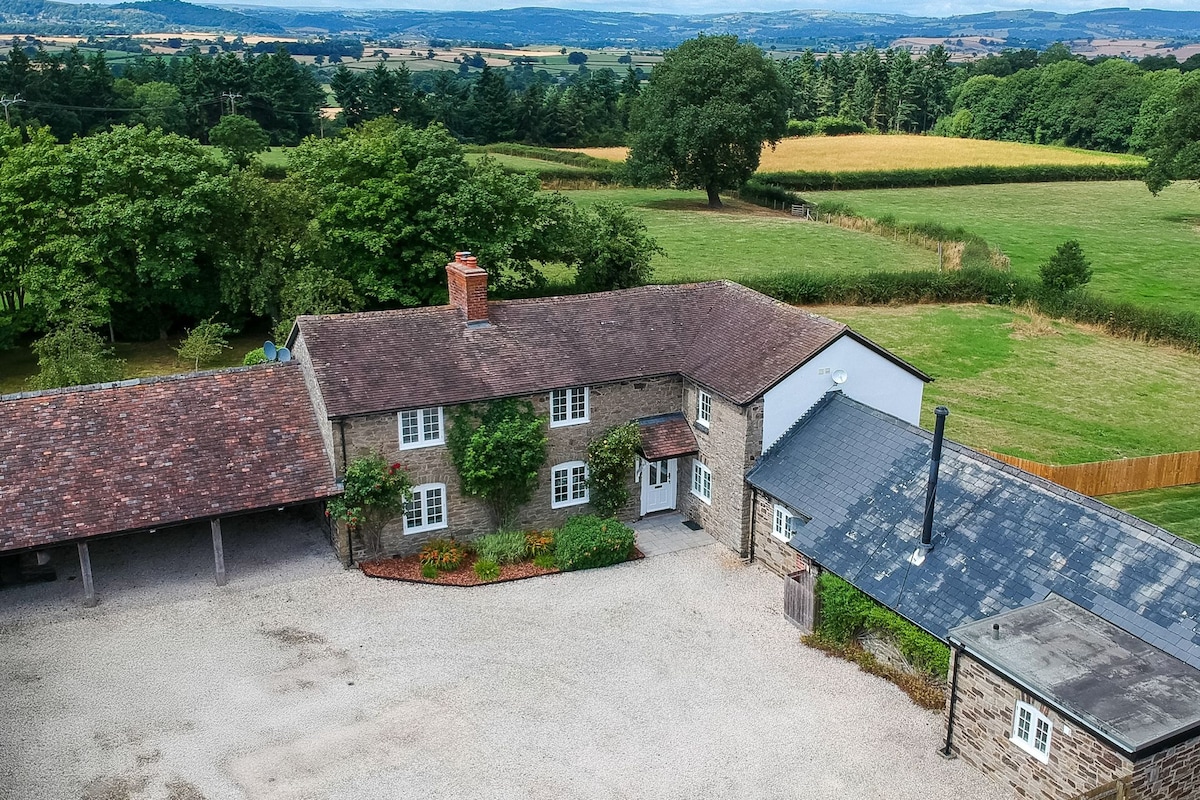 A rural country cottage is set within expansive greenery, featuring a stone exterior and a rustic roof. Surrounding landscapes include rolling fields and trees, offering a serene backdrop. A partially visible gravel driveway leads up to the entrance, hinting at ample outdoor space.