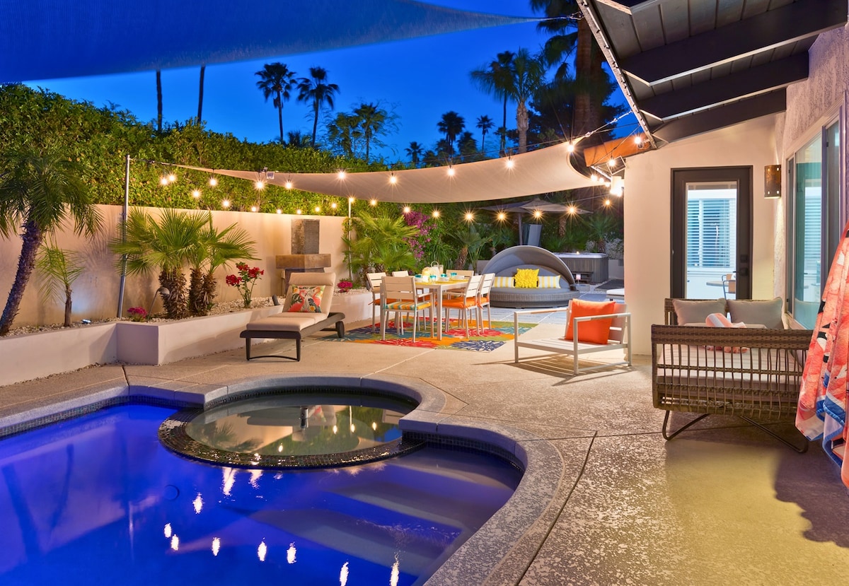 An outdoor living area features a heated saltwater pool and spa, surrounded by lush greenery and palm trees. Colorful furniture includes a dining table and seating area, illuminated by string lights overhead. The space invites relaxation under the night sky, creating a cozy ambiance.