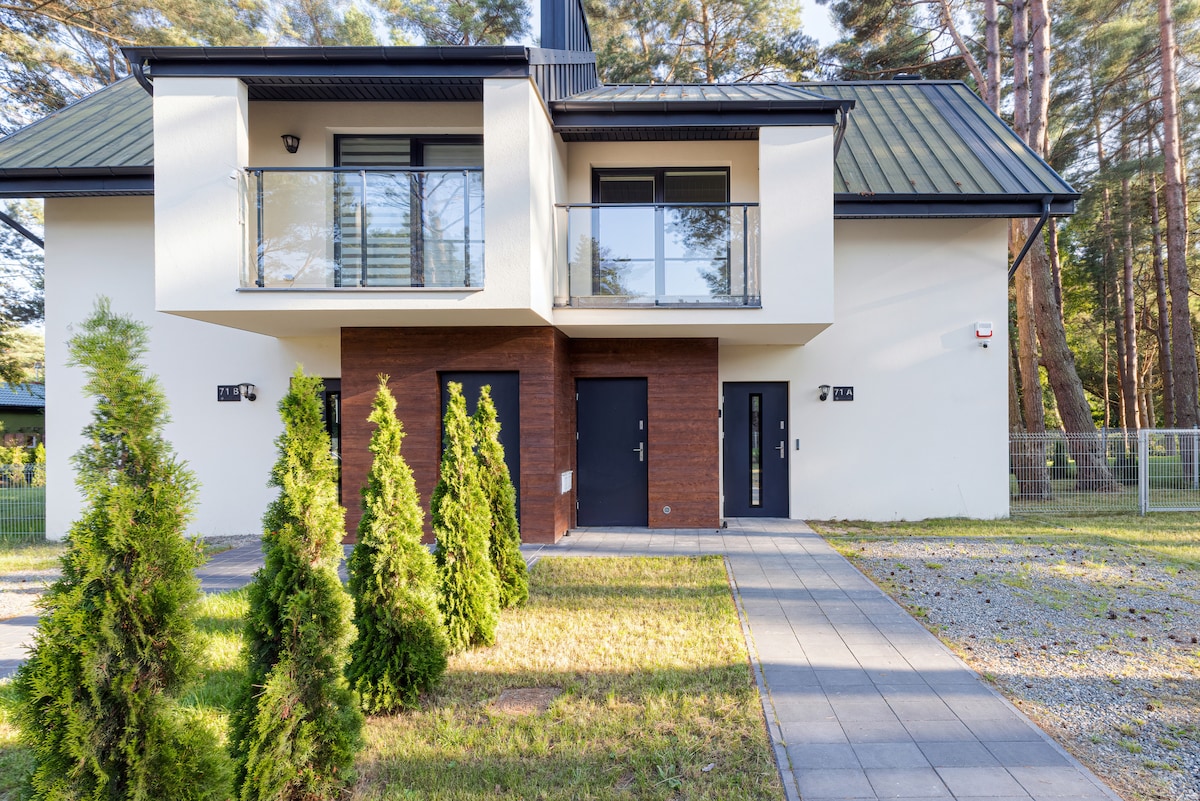 The modern building features a two-story design with a combination of light and dark exterior finishes. Small, decorative trees line the front, and a paved pathway leads to the entrance doors. Large windows allow natural light to flow into the interior.
