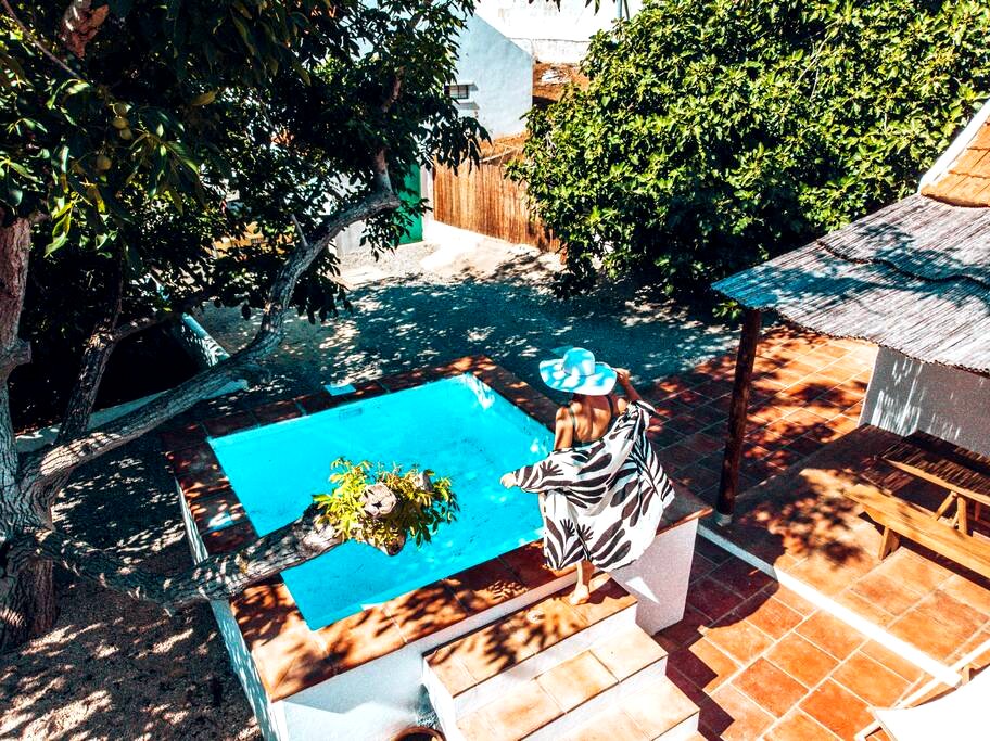 An inviting outdoor area features a private swimming pool surrounded by tiles and shaded by trees. A person relaxes on the edge, draped in a colorful towel. Nearby, a thatched-roof structure provides additional shading, creating a serene space for leisure.