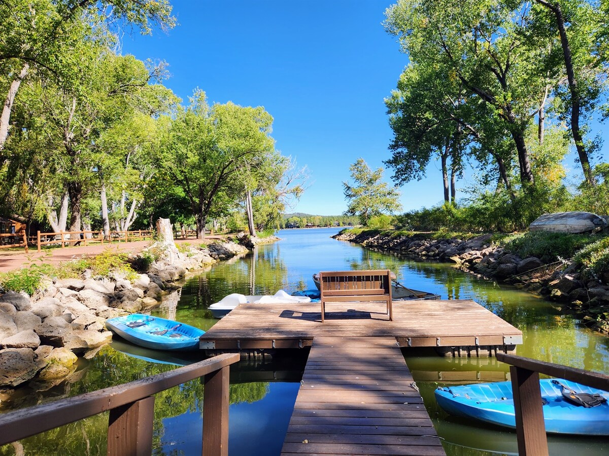 A wooden dock extends over calm waters, surrounded by lush greenery and tall trees. Two kayaks are moored alongside, and a bench is positioned on the dock, offering a serene view of the tranquil lake and natural landscape.