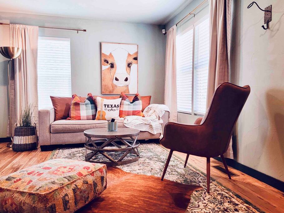 A welcoming living area features a comfortable grey sofa adorned with colorful cushions. A large artwork of a cow is displayed above a round coffee table, complemented by a cozy armchair. Natural light filters through two windows, illuminating the hardwood floor.