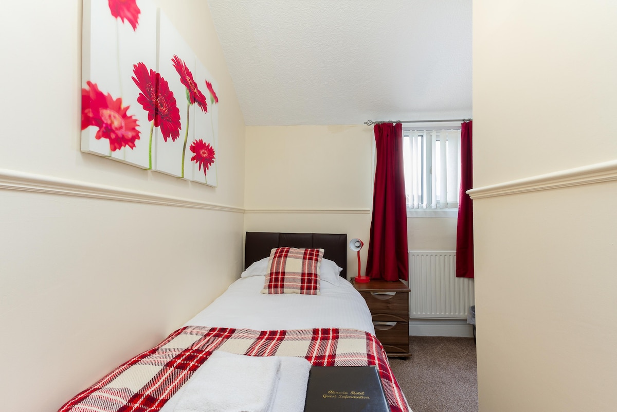 A compact guest room features a single bed adorned with a plaid blanket. Red floral art hangs on the wall above, complementing the red curtains framing a window. A bedside table with a lamp stands next to the bed, along with a folded towel placed on the blanket.