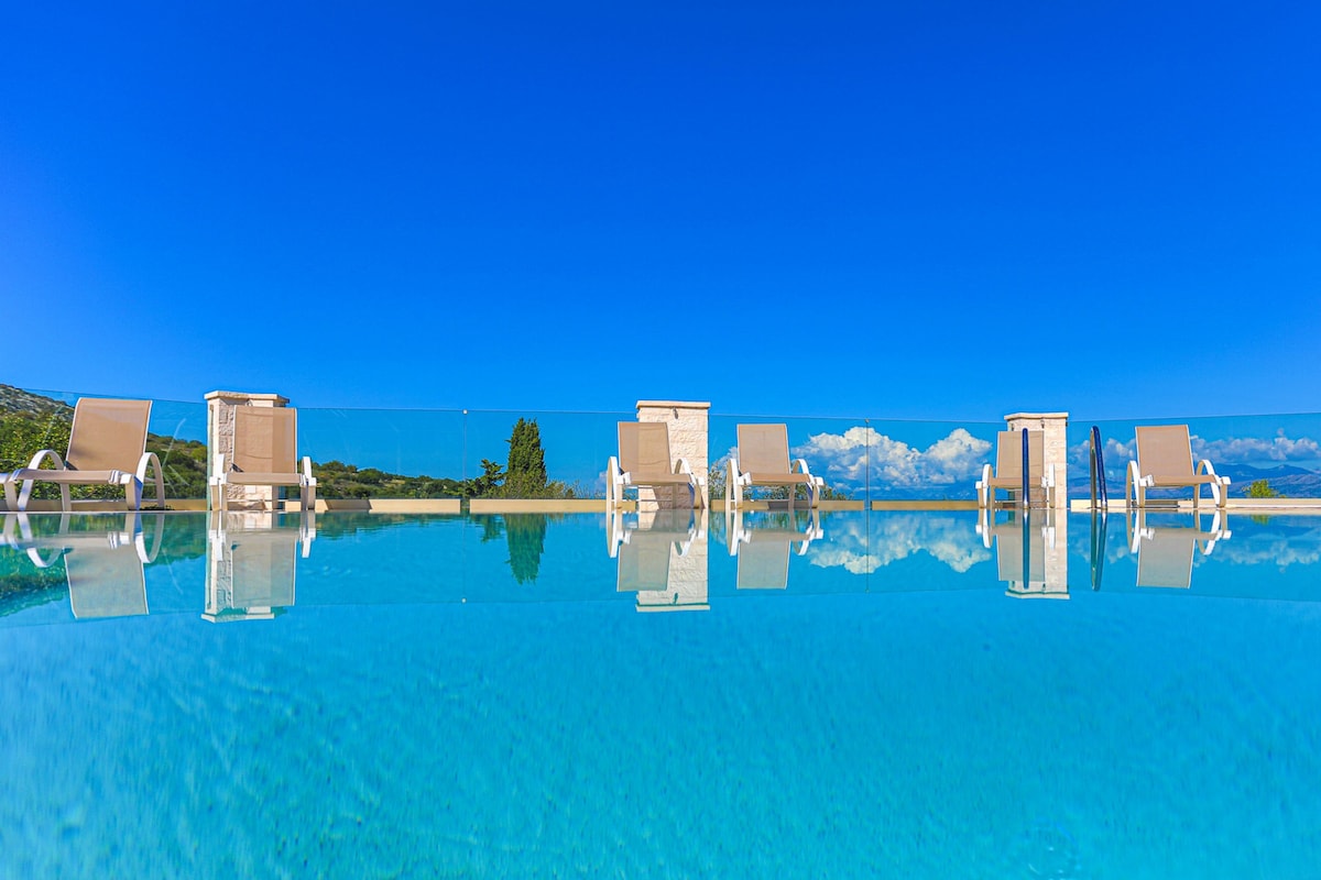 An oval pool with crystal-clear water reflects the surrounding landscape. Sun loungers are arranged along the edge, offering a view of the horizon and distant mountains. The sky above is clear, creating a serene atmosphere.
