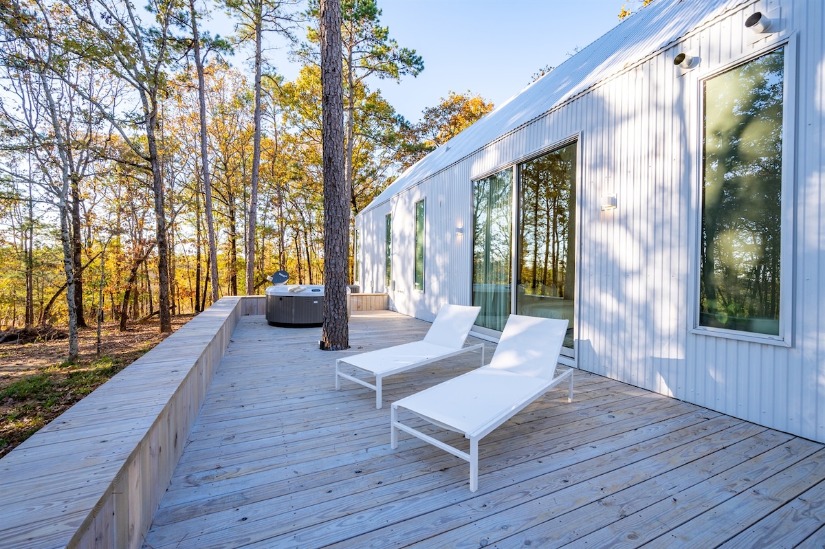 A deck with wooden flooring is furnished with two white lounge chairs, positioned for relaxation. This area is surrounded by tall trees, allowing natural light to filter through, creating a serene outdoor space for unwinding.