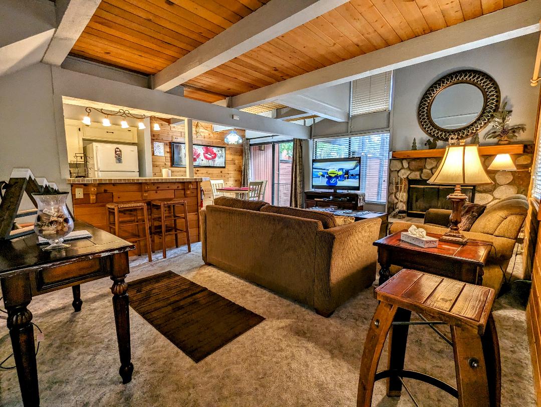 The living area is equipped with a comfortable sofa and armchair set, facing a flat-screen TV. A stone fireplace adds a cozy touch, while light streams through the windows. A dining area with barstools and a wooden table completes the spacious layout.