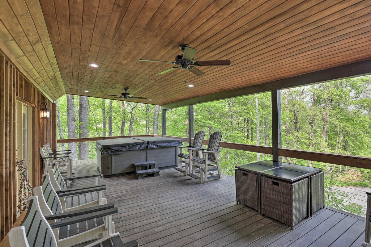 A covered deck area features a private hot tub, comfortable seating with multiple chairs, and a storage box. The wooden ceiling enhances the warm atmosphere, while views of the surrounding greenery can be seen through the railing.