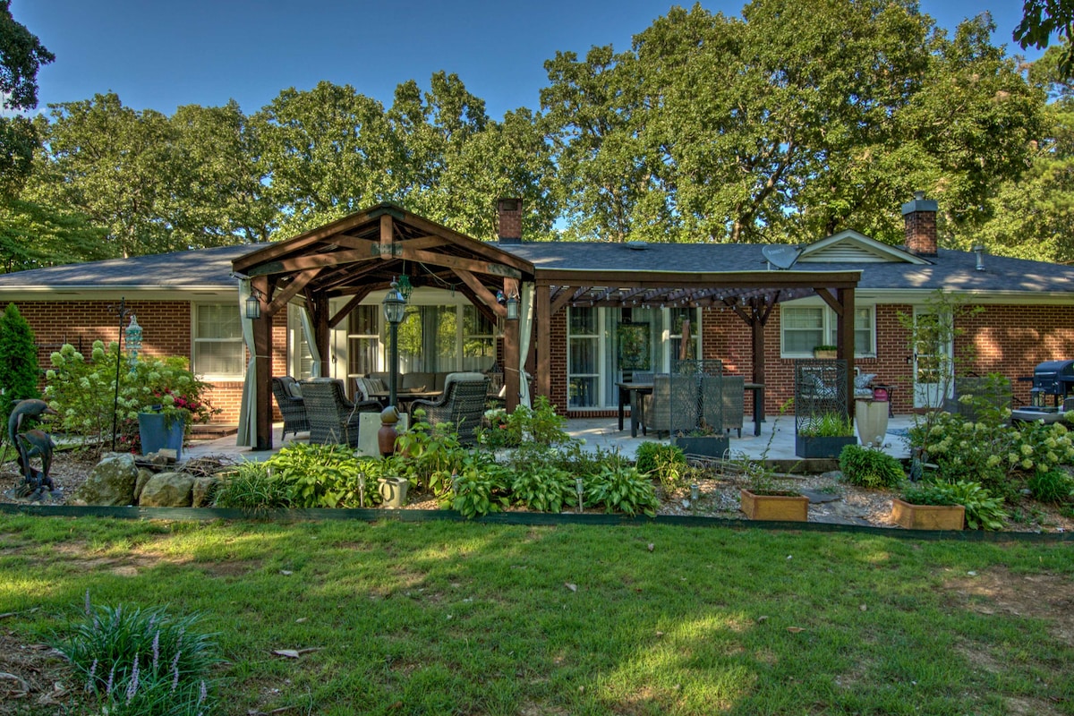 A spacious patio area features a wooden pergola with comfortable seating beneath. Lush greenery surrounds the space, complemented by well-maintained flower beds. A dining table is visible, along with a charcoal grill, creating an inviting outdoor environment.