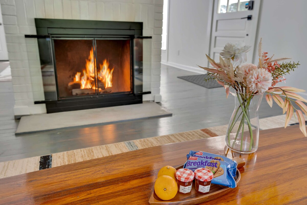 A seasonal wood-burning fireplace is centered within the image, with flames flickering. A wooden coffee table features a small vase of flowers and a snack arrangement, including two oranges and packaged breakfast items. The surrounding floor is light-colored with a flat texture.
