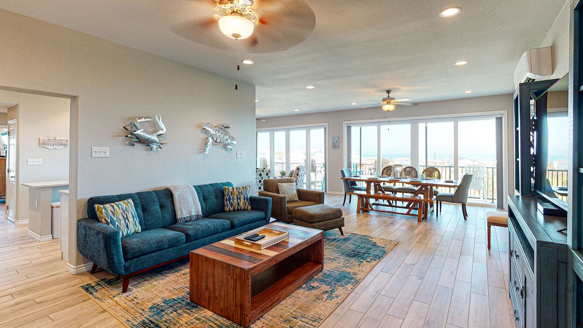 A spacious living area features a dark blue sofa and two accent chairs arranged around a wooden coffee table. Large sliding glass doors provide views of the balcony, while a dining table accommodates ten guests. Decorative fish wall art adds a coastal touch.