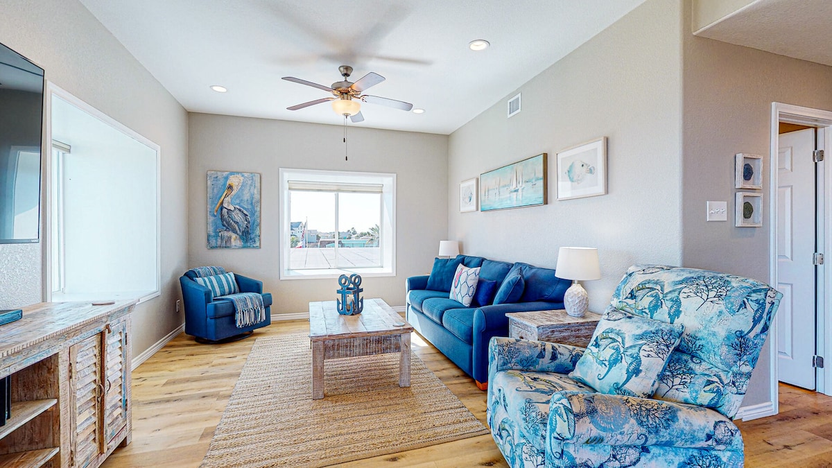A bright living area is furnished with a large sectional sofa and two accent chairs. A coffee table sits in the center, with decorative artwork adorning the walls. Natural light enters through a large window, and a ceiling fan adds comfort to the space.