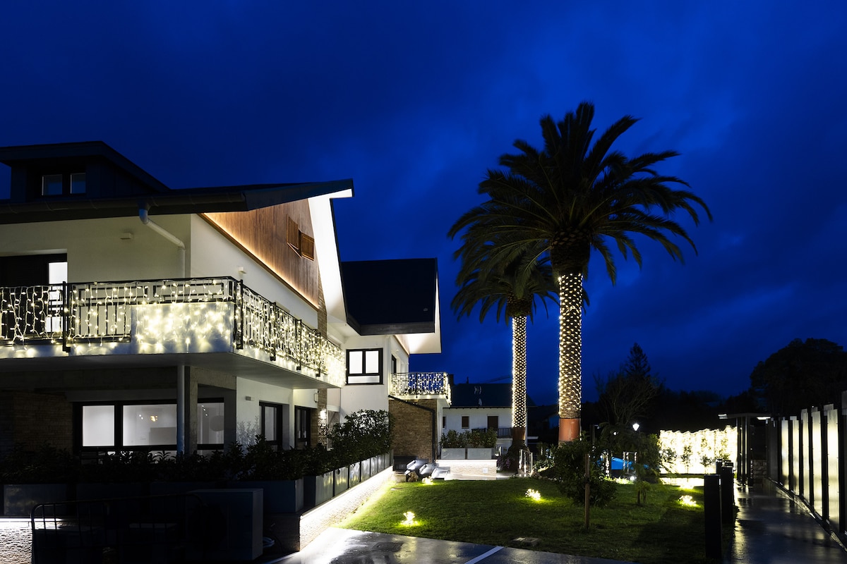 The elegant exterior of the apartment complex is illuminated by soft lighting, highlighting the architectural features. Two tall palm trees are positioned beside the building, surrounded by a landscaped garden. The night sky adds a serene backdrop to the tranquil setting.