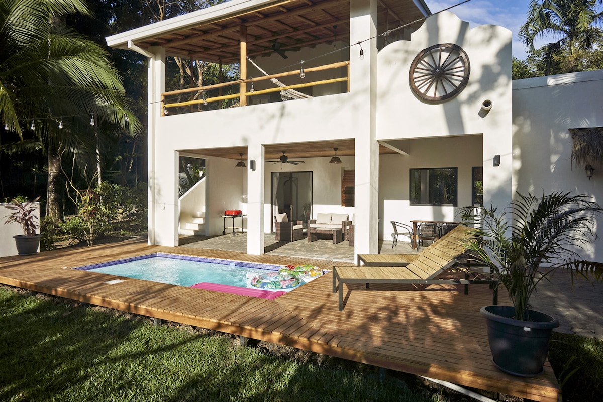 The exterior of the villa features a spacious wooden deck surrounding a private pool. Outdoor seating areas are visible, with a mix of lounge chairs and tables. Tropical greenery lines the property, enhancing the serene environment. Large glass doors suggest access to the interior spaces.