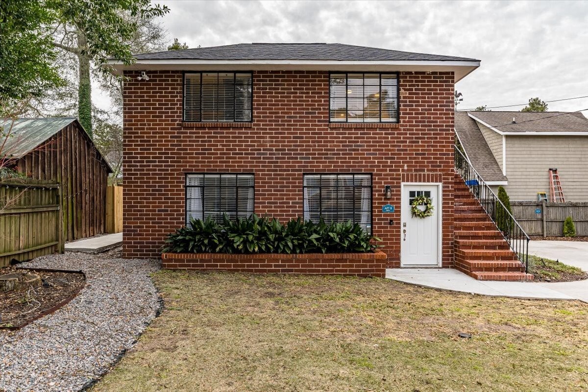 A two-story brick building is showcased, featuring large windows with black shutters. A small set of stairs leads to a white front door with a decorative wreath. The surrounding area is landscaped with green foliage and gravel pathways, providing a welcoming entrance.