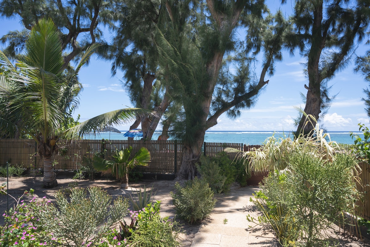 The garden area features lush greenery and tropical plants, enhancing the natural surroundings. A view of the lagoon is visible through tall trees, inviting a sense of tranquility. A sandy path leads towards a fence that borders the beach.