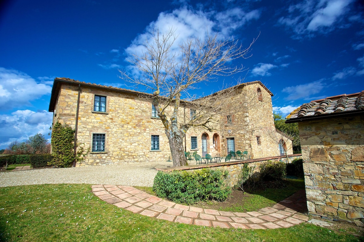 The stone building features multiple windows and a welcoming entryway with arched doorways. A grassy area surrounds the structure, punctuated by a bare tree and a pathway of stone tiles leading to a seating area with green chairs.