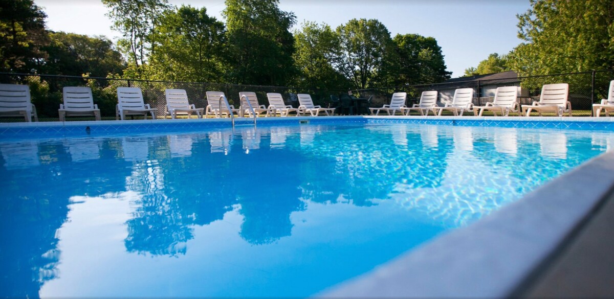 A large outdoor pool is surrounded by multiple lounge chairs, offering ample space for relaxation. Clear blue waters reflect the trees and sky above, creating a serene environment for guests to enjoy. The area is well-lit, ideal for daytime leisure.