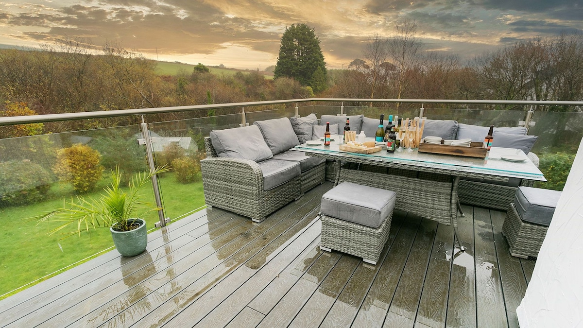 A glass-fronted balcony features a large outdoor dining table surrounded by cushioned seating, designed for relaxation. The wet wooden deck reflects the surrounding greenery, with a small potted plant adding a touch of nature. The sunset casts a warm glow over the tranquil setting.