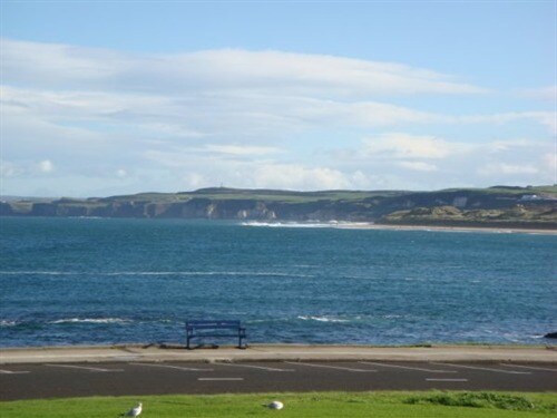 The image shows a tranquil view of the Atlantic Ocean, displaying calm waters and gentle waves. In the distance, cliffs rise against a backdrop of green hills under a partly cloudy sky. A bench is positioned along the waterfront, inviting relaxation and contemplation.