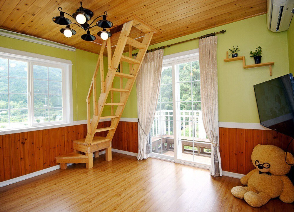 A wooden staircase leads to an upper level in a room featuring light green walls and warm wooden accents. Large windows allow natural light to fill the space, while a cozy seating area is complemented by a large stuffed bear.