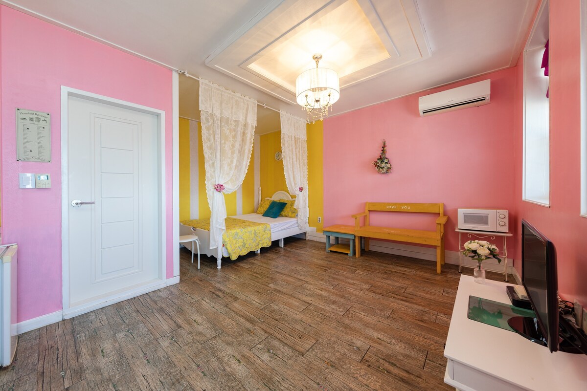 A bright and cheerful room features pink walls and wooden flooring. A single bed is adorned with light curtains, while a yellow seating area adds a pop of color. A microwave and a television are conveniently placed within the space.