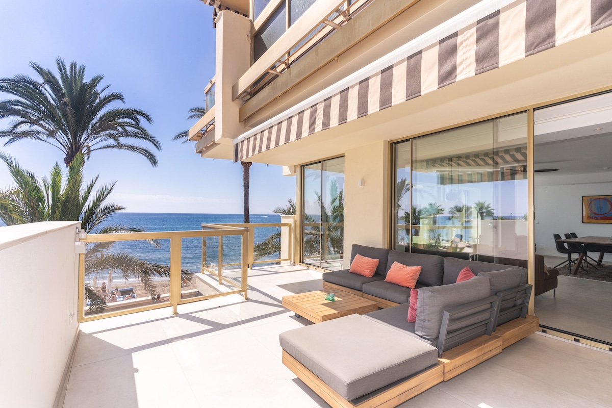 An outdoor terrace features comfortable seating arrangements with plush cushions and a wooden coffee table. Palm trees are visible in the background, framing a serene view of the Mediterranean Sea.