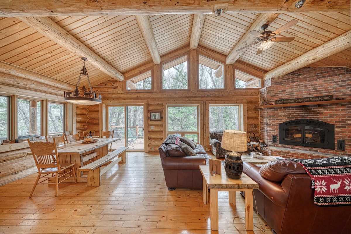The living area features a vaulted wooden ceiling and large windows that allow natural light into the space. A rustic brick fireplace serves as a focal point, while a wooden dining table and comfortable seating arrangements create an inviting atmosphere for relaxation.