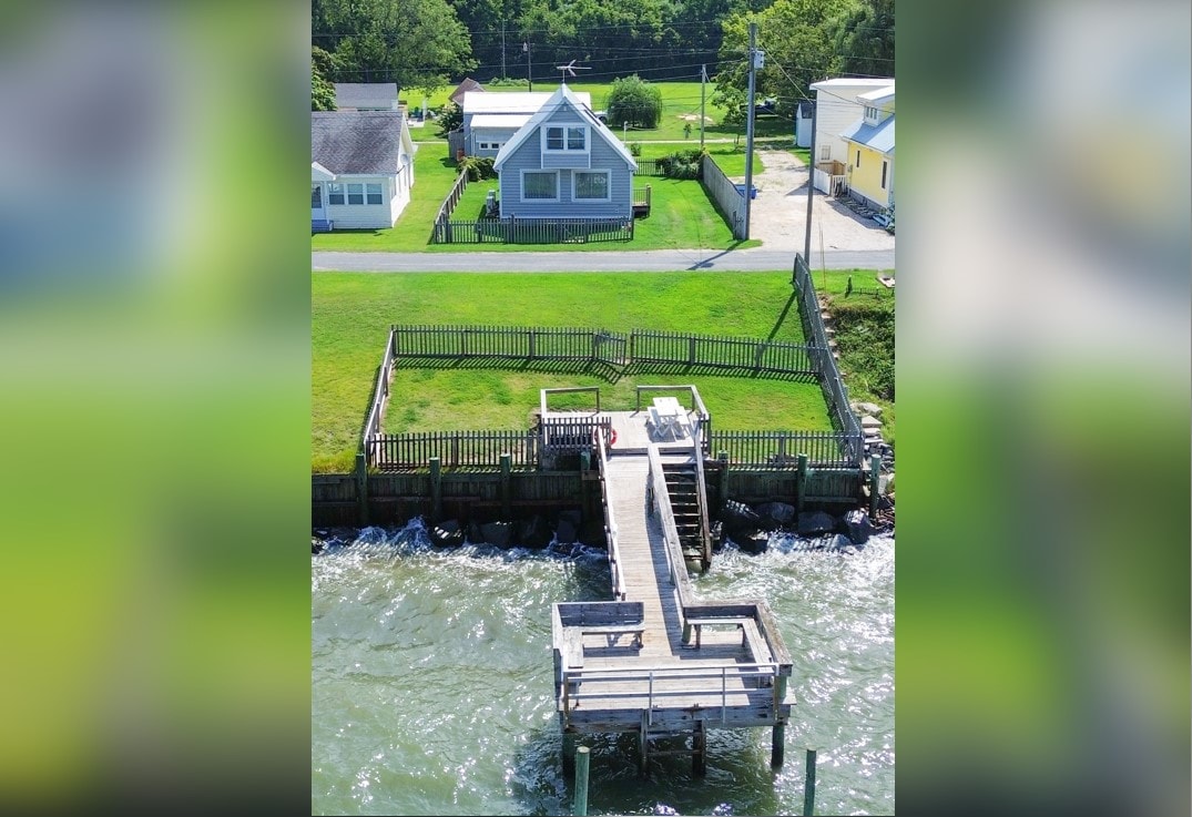Aerial view of the cottage and fenced-in private dock set on the Chesapeake Bay. The dock extends over shallow water, surrounded by green grass. Neighboring homes are visible in the background, showcasing a peaceful waterfront community.