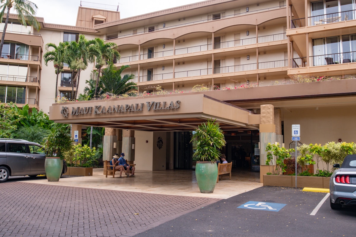 The entrance of Maui Ka’anapali Villas is framed by tropical landscaping, with large potted plants placed at the entrance. The building features a subtle architectural design with multiple floors visible in the background. Seating is provided for guests in front of the entrance.