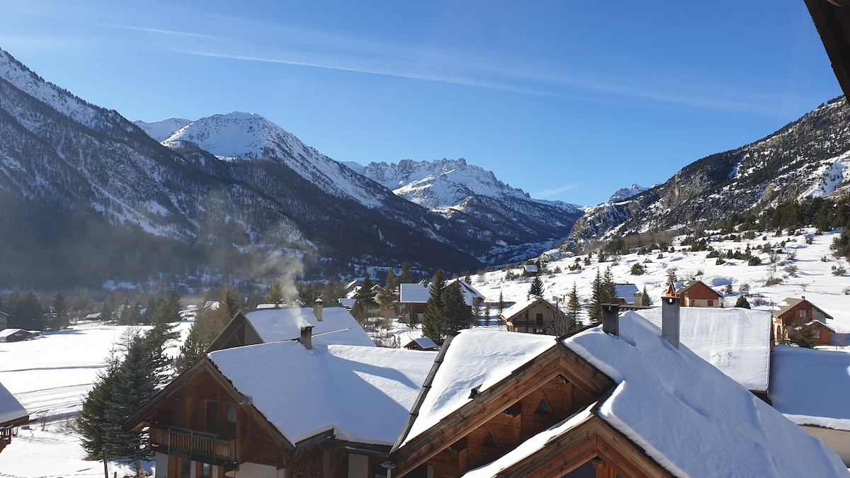 A scenic winter landscape features snow-covered rooftops and expansive views of distant mountains. Clear blue skies stretch above, and the sun casts a gentle glow over the serene valley, enhancing the tranquility of the surrounding countryside.