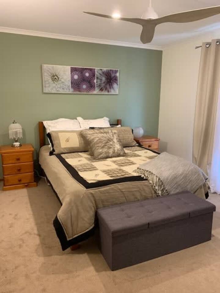 تحتوي غرفة النوم المريحة هذه على سرير متوسط الحجم مع لحاف محايد اللون ووسائد مزخرفة، مقابل جدار أخضر يميل إلى الرمادي. تحيط طاولات خشبية متطابقة مع مصابيح بالسرير، بينما توجد أريكة رمادية ناعمة عند طرفه. الغرفة جيدة