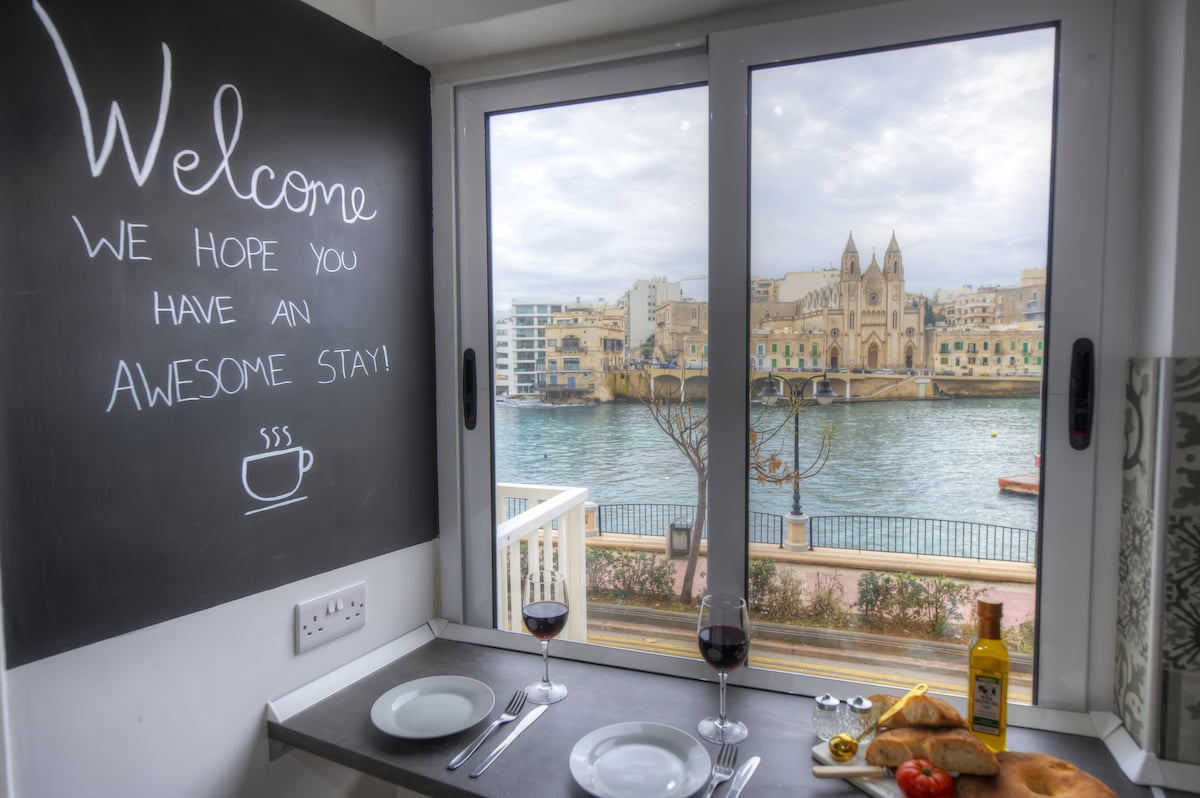 A dining area includes two white plates, wine glasses filled with red wine, and a bottle of olive oil. A chalkboard displaying a welcoming message is positioned next to the window, which offers a view of Balluta Bay and the ornate Balluta Church.