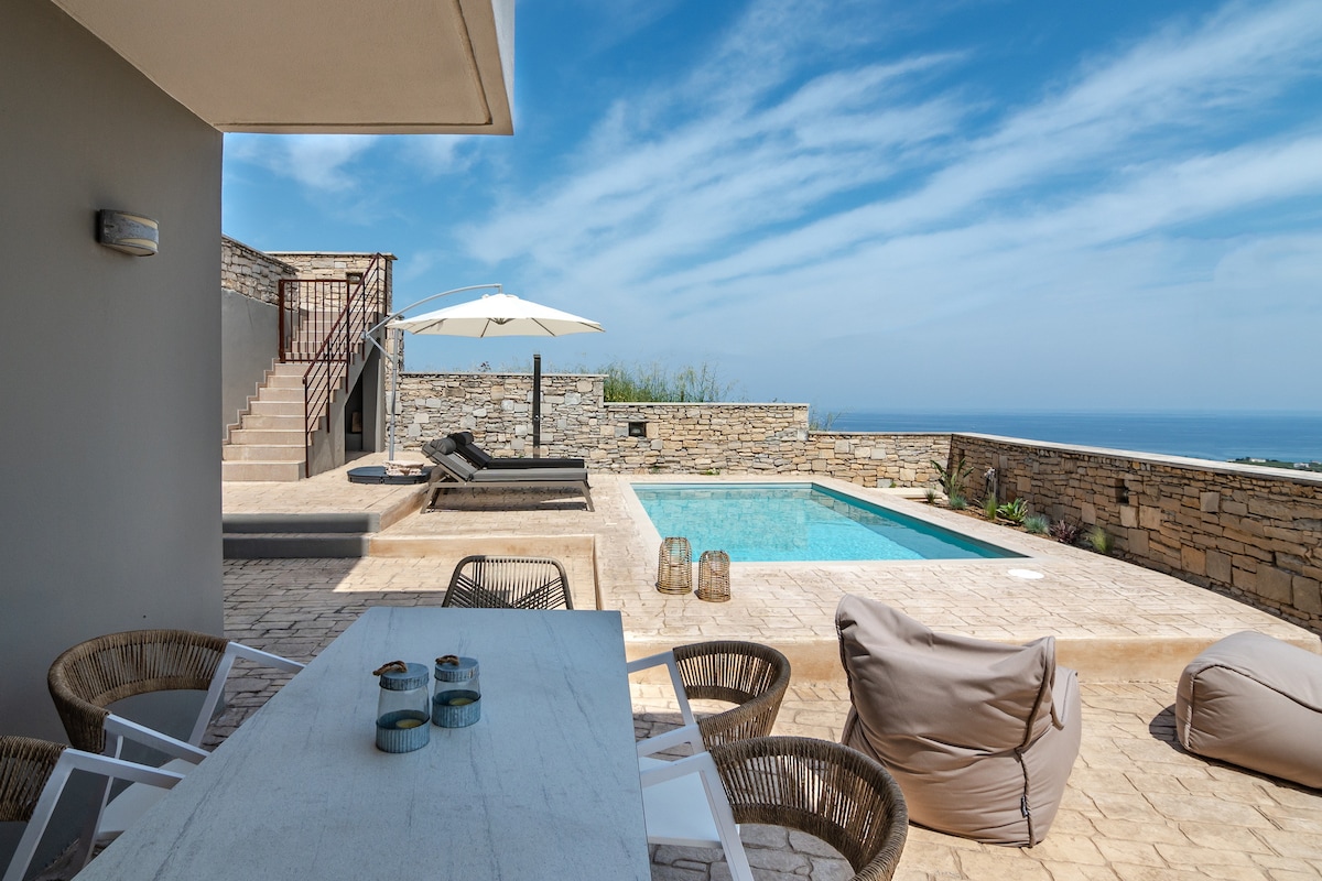 An outdoor terrace features a dining table surrounded by several chairs, with a view of a private pool. A sun umbrella and lounge chairs are positioned nearby, while stone walls enhance the space's natural setting with distant sea views under a clear blue sky.