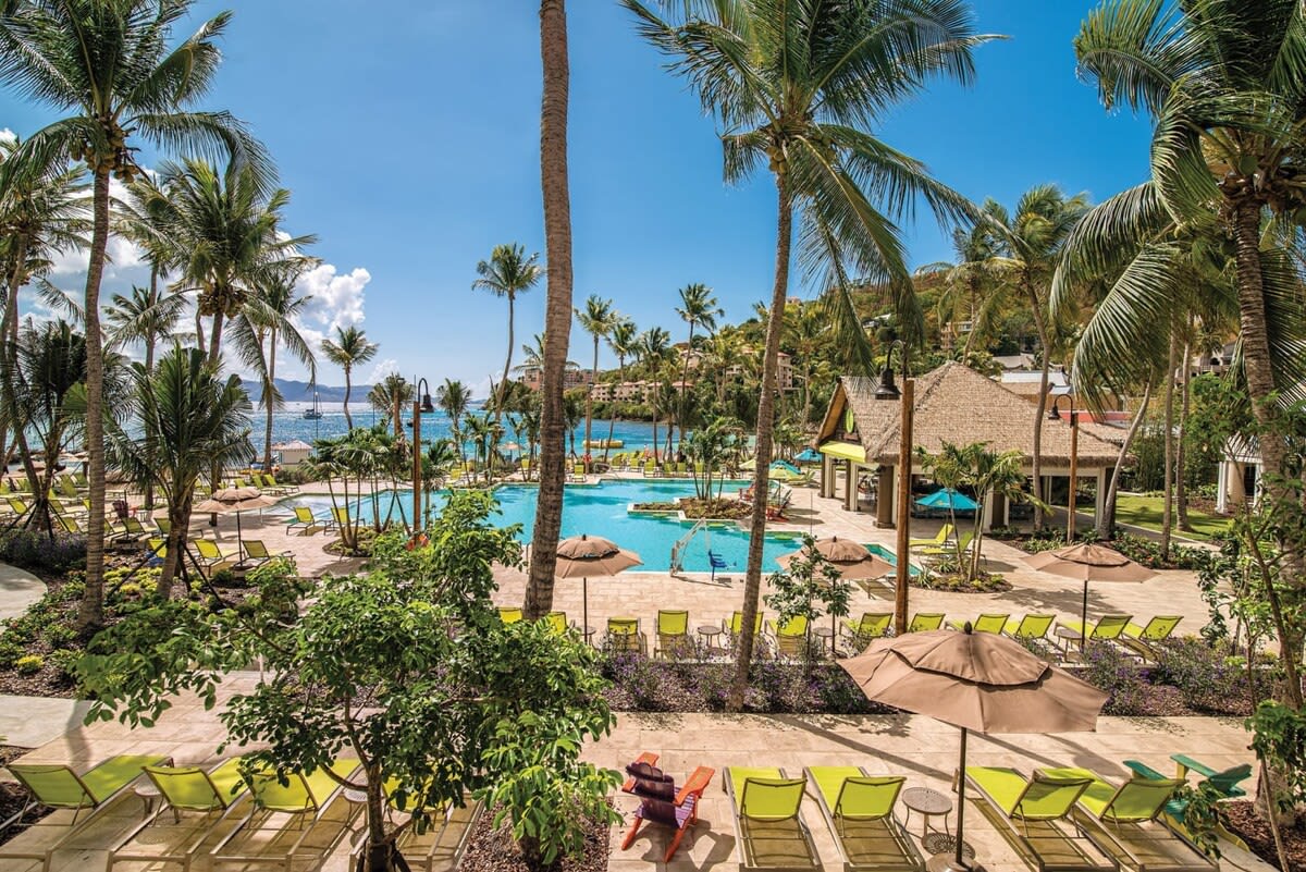 An expansive resort area is shown, featuring a large swimming pool surrounded by palm trees. Comfortable lounge chairs in bright colors are positioned around the pool. A thatched-roof structure can be seen in the background, enhancing the tropical setting with clear blue skies above.
